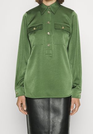 Kvinde iført en grøn skjorte med knapper og klaplommer samt et sort lædernederdel, stående foran en hvid baggrund.