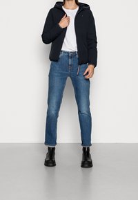 Sort polstret jakke med hætte, hvid t-shirt, blå jeans og sorte ankelstøvler. Jakken har elastiske manchetter og logodetaljer.