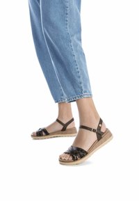 Sandali marroni e neri con motivo, con zeppa bassa, caratterizzati da una fascia alla caviglia e un design incrociato. Abbinati a jeans a tre quarti di colore blu chiaro.