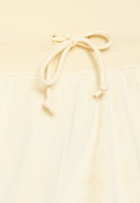 Krémová látka s jemnou texturou, hladkým horním dílem a nařaseným spodním dílem. V pase poutko se šňůrkou a ozdobným uzlem.