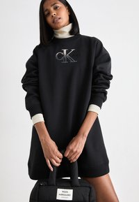 Svart oversized sweatshirt med rund hals, har en silverfärgad präglad logotyp, ribbade ärmslut och en mjuk textur, ihop med en svart quiltad väska.