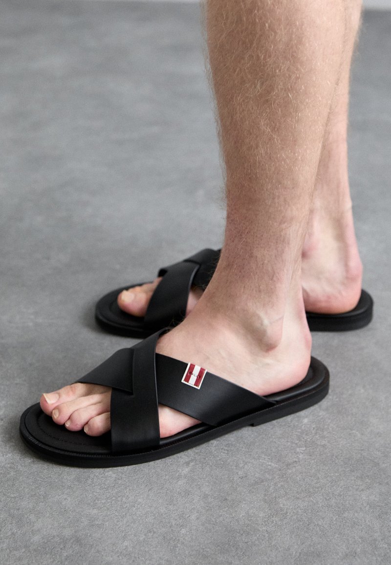 Black sandals with crossed straps, made of smooth material. Features a small red and white logo detail on the side. Flat sole design.