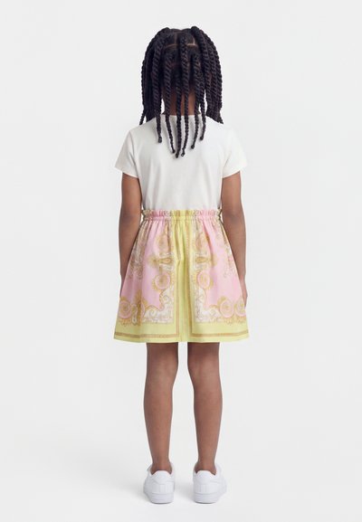 Παιδί που στέκεται με την πλάτη προς τα εμπρός, φοράει λευκό κοντομάνικο μπλουζάκι, φούστα με ροζ και κίτρινο μοτίβο, λευκά αθλητικά παπούτσια και έχει τα μαλλιά του πλεξούδες.