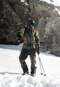 L'attrezzatura per gli sport invernali comprende una giacca impermeabile verde scuro e pantaloni, guanti neri e bastoncini da sci. Una tavola da snowboard è appoggiata sulla spalla.