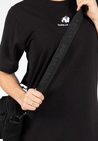 Zwarte katoenen t-shirt met geborduurd wit logo, vergezeld van een zwarte canvas tas met een gestructureerde, verstelbare band met branding.