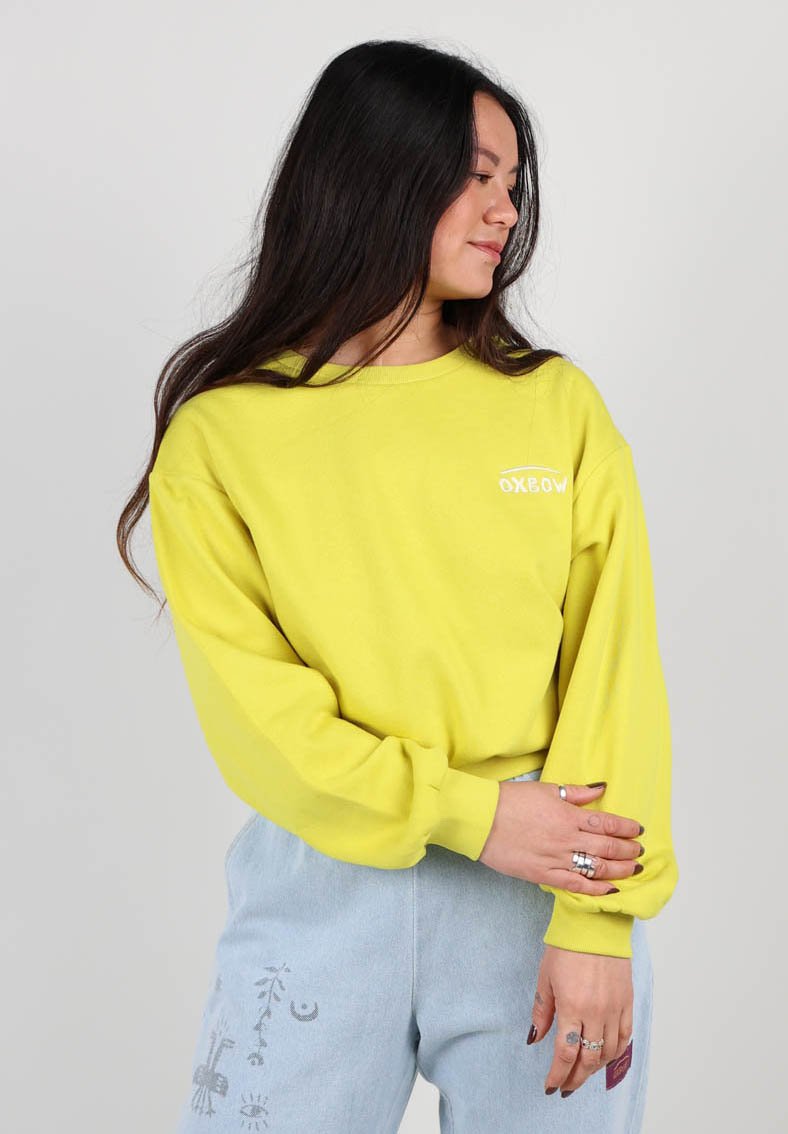 Sweatshirt jaune à manches longues avec un col rond et une texture douce, arborant un petit logo blanc sur la poitrine. Associé à un pantalon bleu clair.
