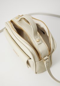 Sac en cuir couleur crème avec fermeture éclair et quincaillerie dorées, doté d'une poignée supérieure et d'une bandoulière ajustable, présentant une finition texturée.