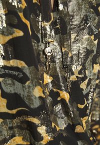 Gros plan d'un tissu avec un imprimé camouflage noir, or et vert, comprenant des fils métalliques brillants et deux tirettes de fermeture éclair argentées avec des breloques en forme de pointes.
