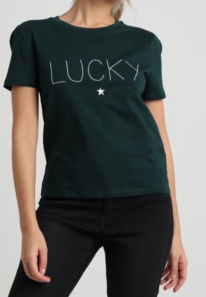 Kvinde iført mørkegrøn T-shirt med teksten "LUCKY" og en hvid stjerne nedenunder, kombineret med sorte bukser mod en ensfarvet baggrund.