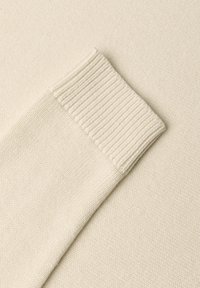 Manche de pull en maille couleur crème avec bord-côtes, posé à plat sur un tissu assorti.