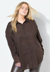 Camisa marrón abotonada con detalles de encaje en las mangas, con un corte holgado y mangas largas. Textura suave, diseño de cuello clásico.