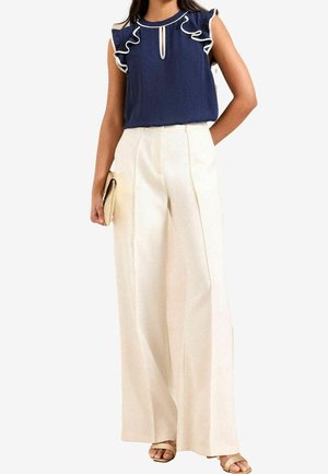 Kvinde iført navy ærmeløs bluse med hvide flæser og højtaljede, vidde ben i cremefarvet bukser, der holder en beige clutch.