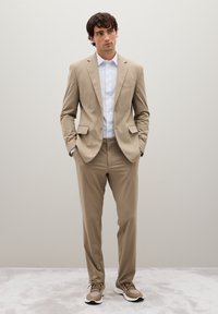 Completo beige slim-fit realizzato in tessuto liscio, con blazer a un petto e pantaloni affusolati, abbinato a una camicia bianca e sneakers casual.