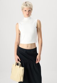 Hvit ermeløs topp med høy krage, kombinert med et samlet svart skjørt, båret med en beige totebag med korte håndtak og lommedetaljer.