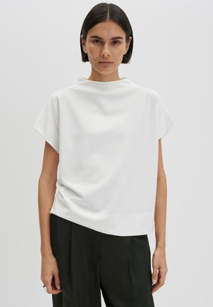 Donna con capelli corti e scuri che indossa una maglia bianca larga e pantaloni neri, in piedi di fronte a uno sfondo chiaro e uniforme.