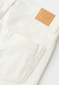 Detail der Gesäßtasche einer weißen Jeans mit sichtbaren Nähten und einem braunen Lederetikett der Marke am Bund.