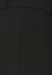 Pantalones de tela negra que muestran una textura suave, un ajuste a medida, detalle de bolsillo lateral y costuras limpias a lo largo de las costuras.