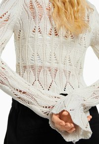 Biały dziergany sweter z ażurowym wzorem diamentów, długimi rękawami i szerokimi mankietami. Tekstura jest luźna i przewiewna.