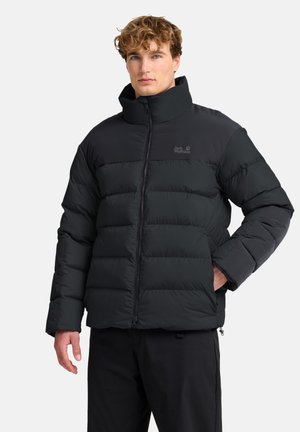 Junger Mann trägt eine schwarze Steppjacke mit hohem Kragen, steht mit einer Hand in der Tasche, neutraler Gesichtsausdruck, einfarbiger Hintergrund.