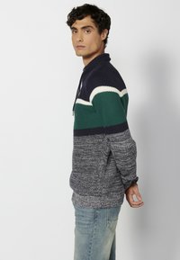 Sweter typu pulower wykonany z dzianiny, z kolorowym wzorem w odcieniach granatu, zieleni i bieli. Rękawy i dół wykończone ściągaczem z teksturowanym wykończeniem.