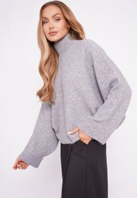 Jasnoszara, oversize sweter z miękkiej, teksturowanej tkaniny, z wysokim prążkowanym dekoltem i szerokimi rękawami; zestawiony z czarnymi spodniami z boczną kieszenią.
