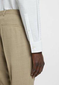 Chemise blanche texturée avec des poignets à boutons, associée à un pantalon beige ajusté avec une poche arrière. Le poignet a un léger brillant et des boutons en perle.