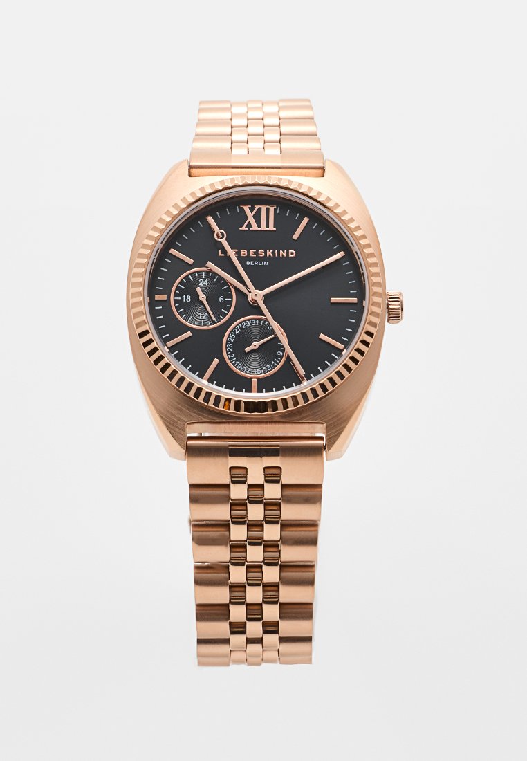 Reloj de pulsera en oro rosa con esfera negra, números romanos y dos subesferas. Presenta una pulsera de metal pulido. Diseño elegante y moderno.