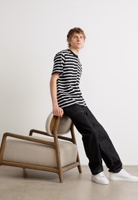 Tricou striped alb și negru, pantaloni închiși și teniși albi. Individul se sprijină de un scaun minimalist din lemn cu tapițerie bej.