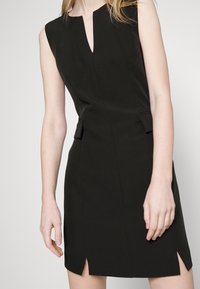 Black sleeveless dress with a V-neckline, side pockets, a fitted design, and subtle front slits, made from smooth fabric.