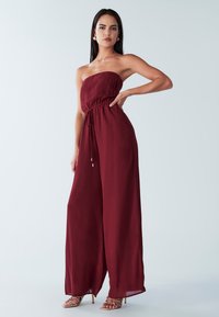 Bordo brez naramnic kombinezon z nabranim zgornjim delom in elastičnim pasom, z širokimi hlačnicami in gladko, lahko tkanino. Sandali z zlatimi poudarki.