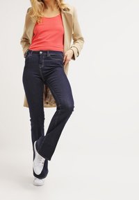Jeans svasati a vita alta di un blu scuro con una texture liscia, abbinati a un top senza maniche rosa e a un cappotto beige, completati da sneakers bianche.