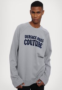 Šedá mikina s rebrovaným okrajom krku a dlhými raglánovými rukávmi. Na prednej strane sa nachádza veľké námornícke modré písmo "VERSACE JEANS COUTURE". Mäkčený materiál.