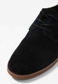 Chaussure en suède noire avec un bout plat, des lacets ronds et une semelle en caoutchouc marron, affichant une texture lisse et un minimalisme dans les détails.