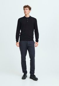 Polo negru cu mâneci lungi și butoane, asortat cu pantaloni închiși la culoare și pantofi negri, pe un model pe un fundal deschis.