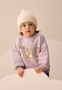 Sweat-shirt violet avec des motifs ludiques, des poignets côtelés et un col montant. Le modèle porte un bonnet en tricot crème. L'arrière-plan présente des couleurs douces et atténuées.