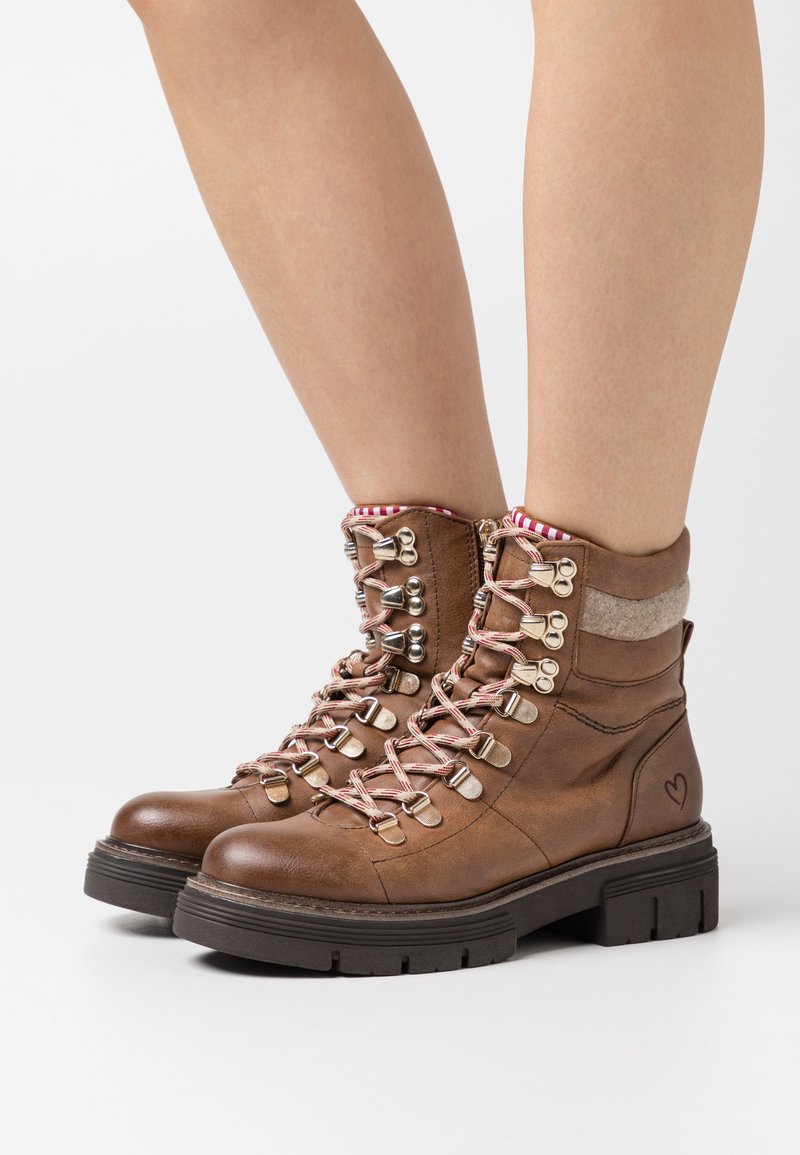 Bottines en cuir marron avec des lacets à motifs rose et blanc, crochets en métal argenté, col rembourré, et un détail en forme de cœur sur le côté. Semelle épaisse noire.