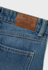 Jeans in denim blu con una etichetta in pelle sul cinturino, caratterizzati da cuciture a contrasto e un dettaglio sulla tasca posteriore.