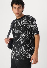 Black textured t-shirt featuring a white abstract print. The model holds a black shoulder bag, with short sleeves and a crew neckline.