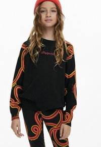 Schwarzer Pullover mit bunten, wirbelnden roten und gelben Mustern an den Ärmeln, ausgestattet mit gesticktem "desigual"-Logo. Kombiniert mit passenden Hosen.