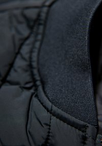 Svart vadderad jacka med en slät yta, som har en kontrasterande vävd krage och synliga sömdetaljer.