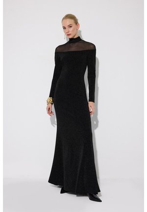 Vrouw in een lange zwarte nauwsluitende jurk met een doorschijnende inzet en een col, gecompleteerd met een grote gouden armband en hangende oorbellen.