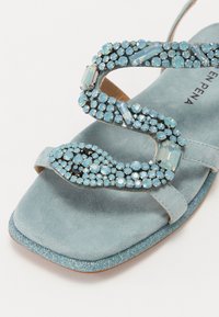 Sandale en suede bleu avec des embellissements en strass bleu clair et noir scintillants. Elle présente un design de lanière ondulée et une semelle plate.