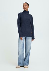 Mørkeblå strikket turtleneck sweater med en tekstureret finish, parret med lyseblå brede jeans og beige slip-on sko.