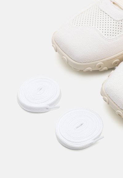Λευκά σουέτ αθλητικά παπούτσια με διάτρητο σχέδιο και μπεζ καουτσούκ σόλα, συνοδευόμενα από δύο τυλιχτά λευκά κορδόνια.