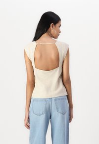 Beige linnedragt med korte ærmer, der har en stor udsparing i ryggen og en lukning i toppen. Stylet med lyseblå, løstsiddende jeans.