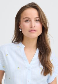 Jeune femme aux longs cheveux bruns portant des boucles d'oreilles argentées et une chemise rayée bleu clair avec de petits imprimés d'ananas, fond neutre.