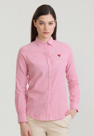 Kobieta o długich ciemnych włosach, ubrana w różowo-białą paskowaną koszulę zapinaną na guziki z małym czerwonym serduszkiem na lewej piersi.