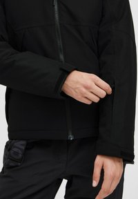 Czarna kurtka z zamkiem błyskawicznym z przodu, charakteryzująca się gładką teksturą, rzepami na mankietach i kieszenią po boku, ukazana z ręką sięgającą po nią.