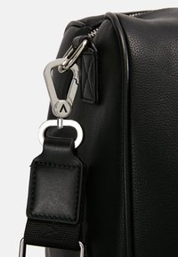 Black synthetic bag detail featuring a silver metal clip, robust stitching, and a textured surface. Strap includes a smooth leather accent.