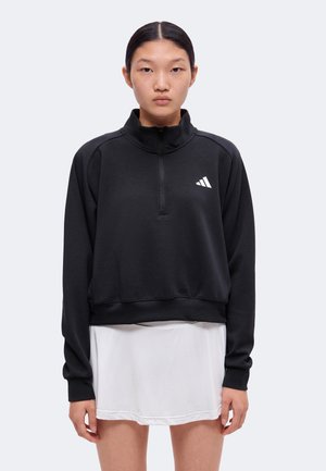 Ung kvinde med sort sweatshirtdragt med lynlås og hvid nederdel står mod en ensfarvet baggrund, vendt fremad med neutralt udtryk.